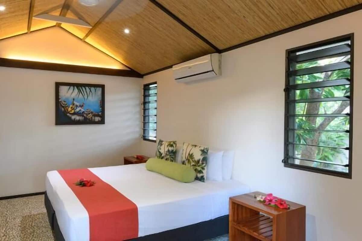 Een slaapkamer met een bed, een schilderij, een airconditioner en een raam met uitzicht op groen.