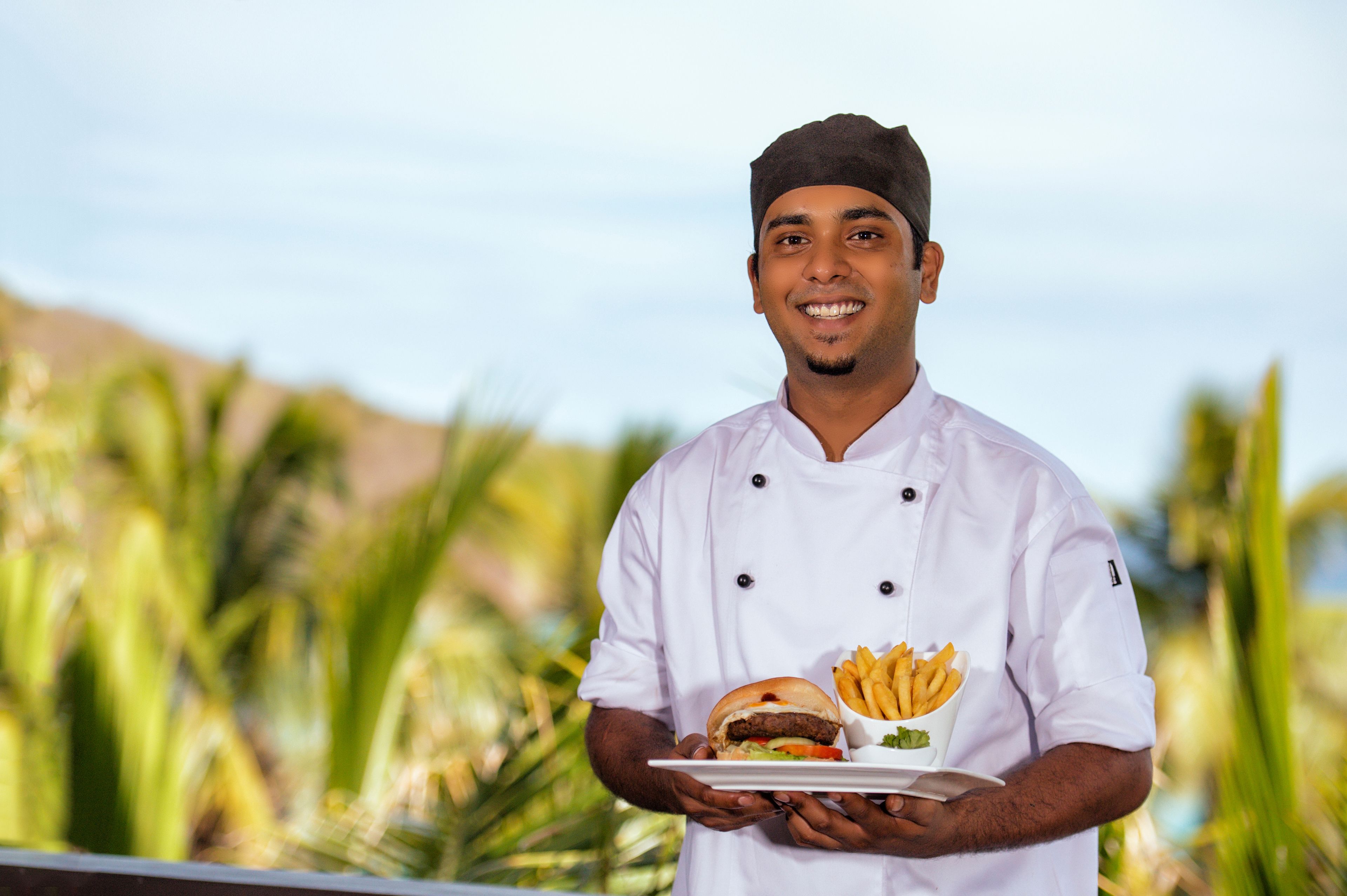 Un chef sostiene un plato con una hamburguesa y papas fritas, de pie al aire libre con palmeras en el fondo.