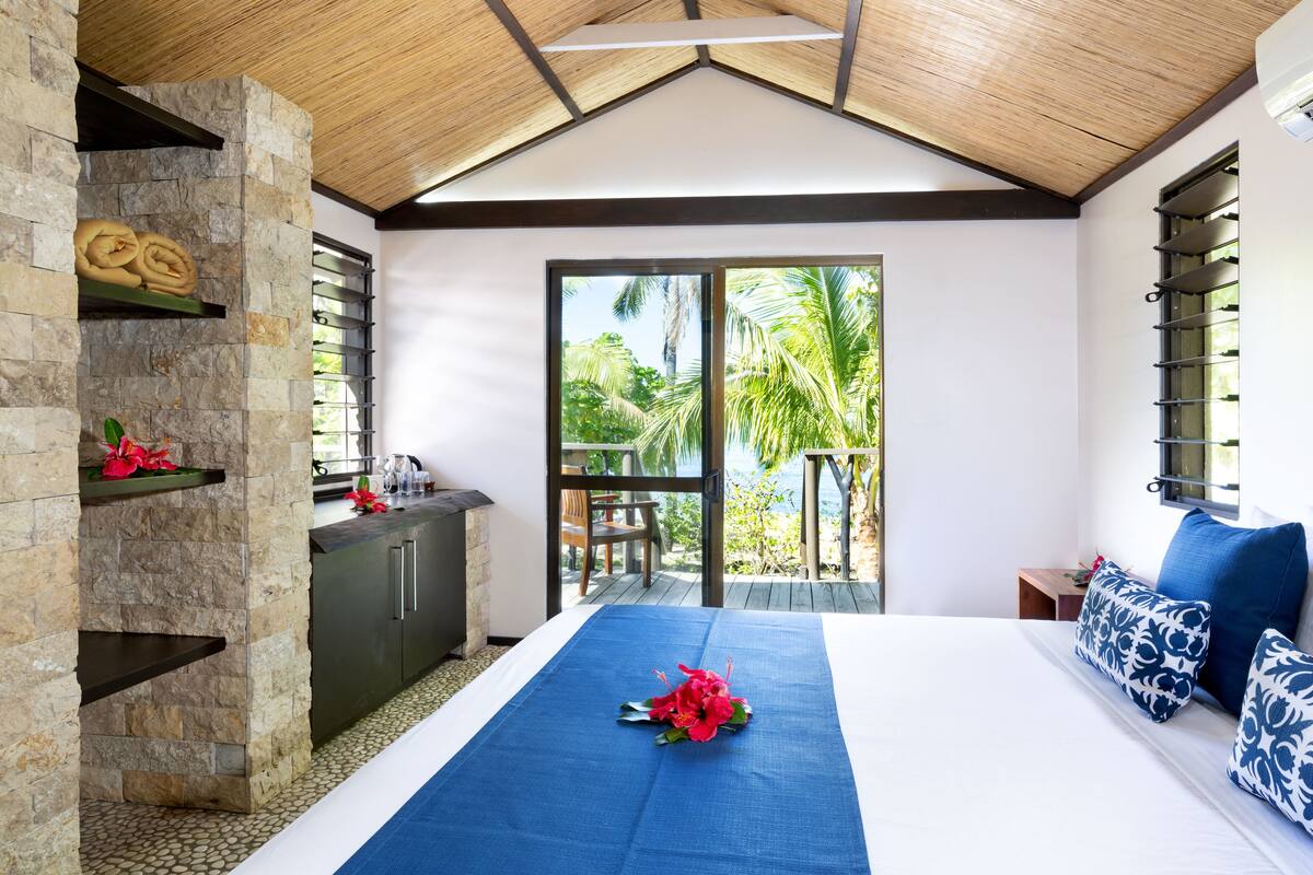 Een hotelkamer met een stenen muur, een bed met een blauwe spreidek en uitzicht op palmbomen door de balkondeuren.
