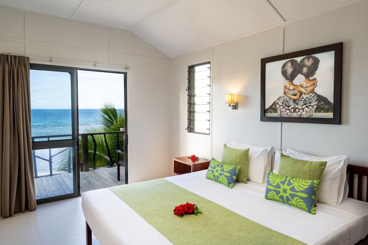 Een hotelkamer met een balkon, een bed met groen en witte beddengoed, een lijst met kunst aan de muur en een rode bloem op het bed.
