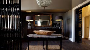 A round table with a wooden bowl centerpiece, flanked by two lamps, in a room with dark wood flooring and a large mirror above.