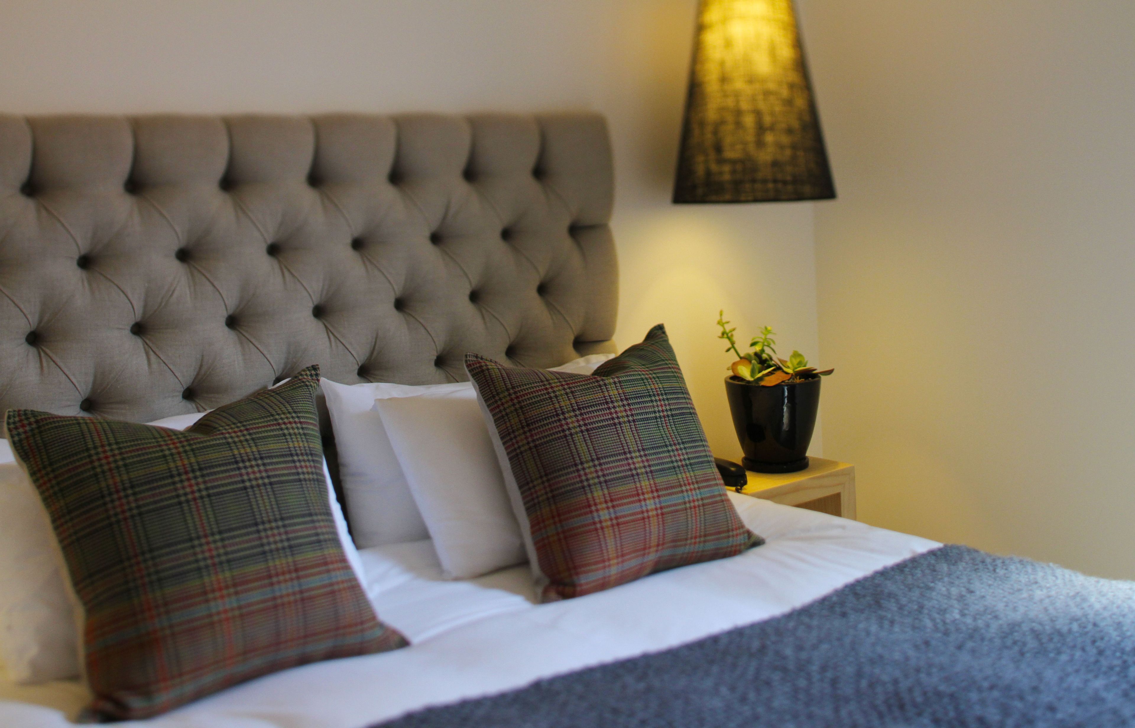 A neatly made bed with a tufted headboard, two plaid pillows, and a bedside table with a lamp and a potted plant.