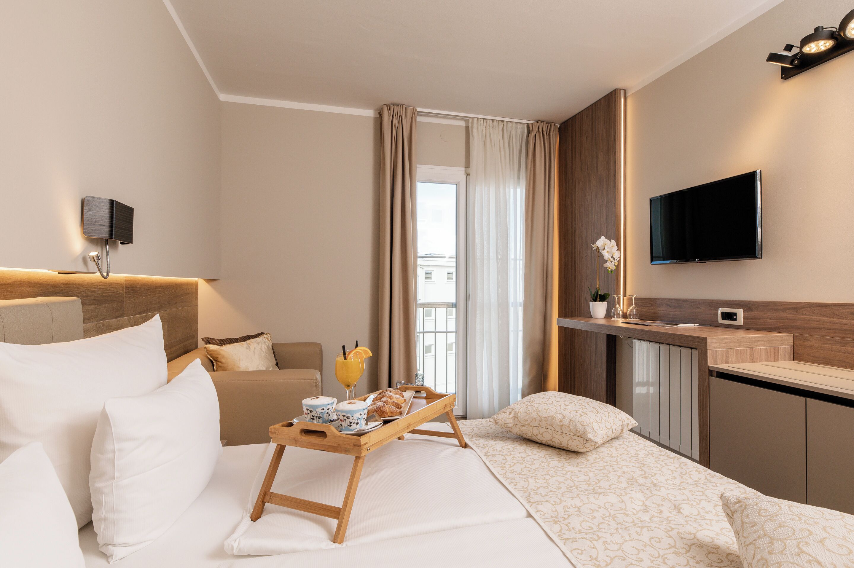 A modern hotel room with a bed, a sofa, a breakfast tray, a TV, and a window with curtains.