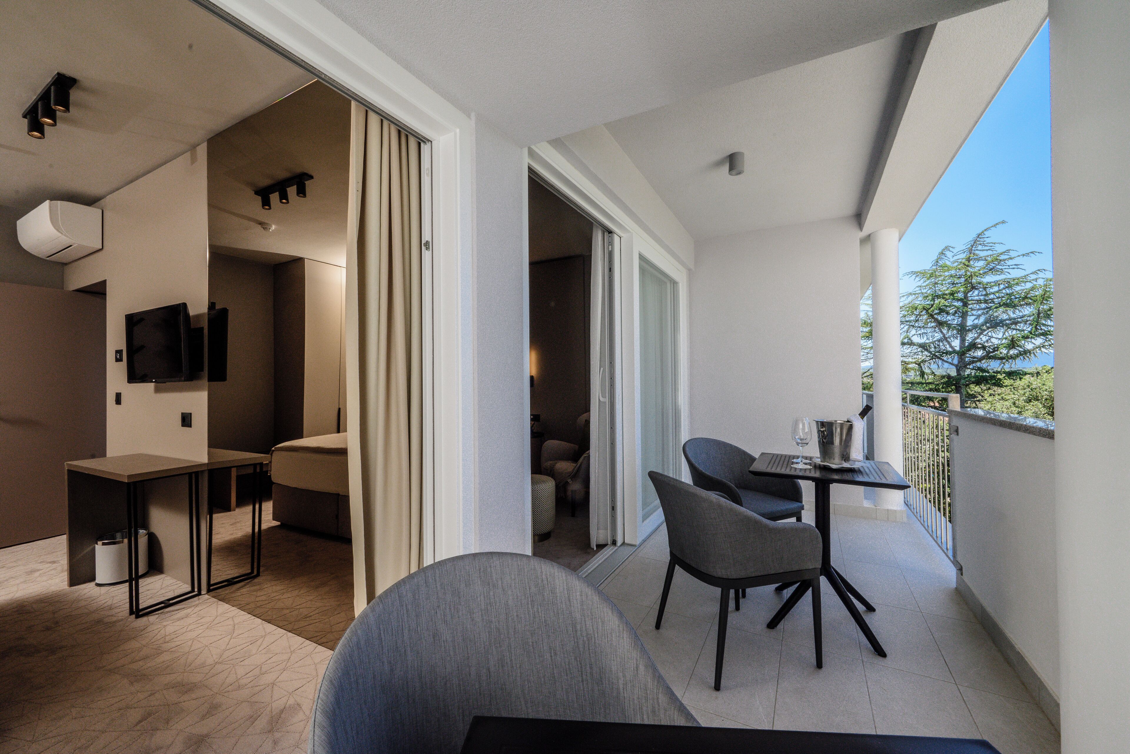 A balcony with a table and chairs, a TV mounted on the wall, and a view of trees.