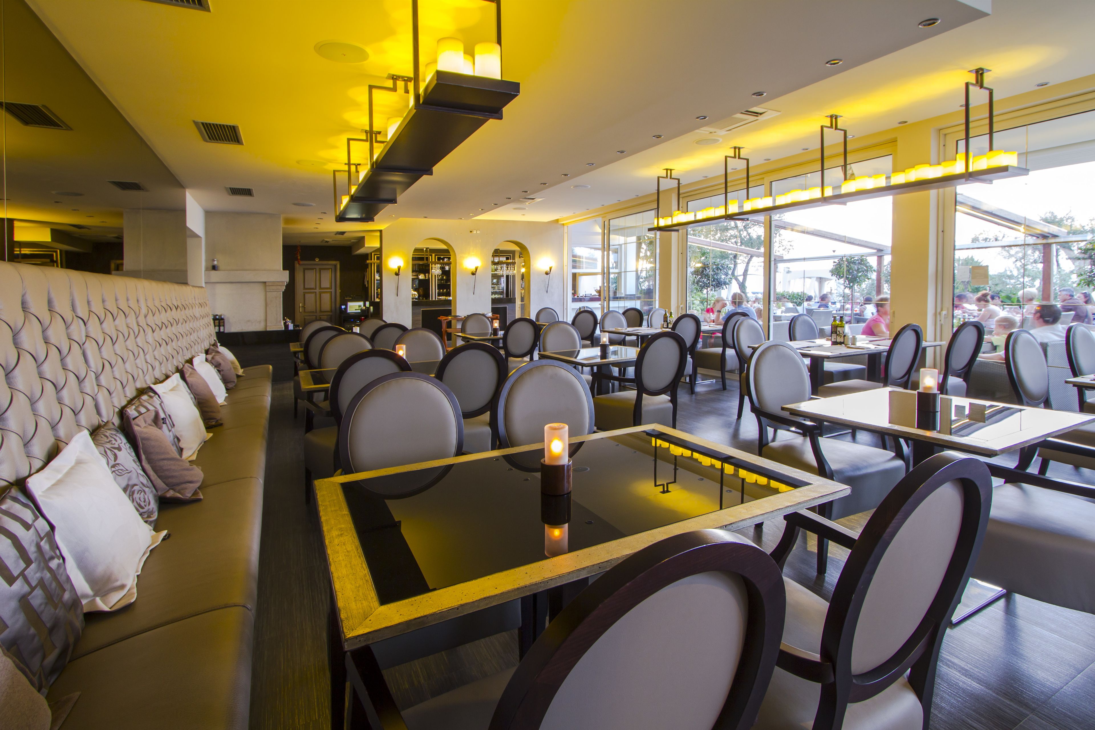 A restaurant with a modern interior, featuring a long bench seating area, tables with chairs, and large windows allowing natural light.