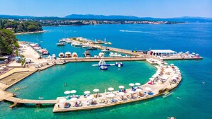 Vista aérea de un puerto deportivo costero con barcos, un muelle y una zona de playa con tumbonas y sombrillas.