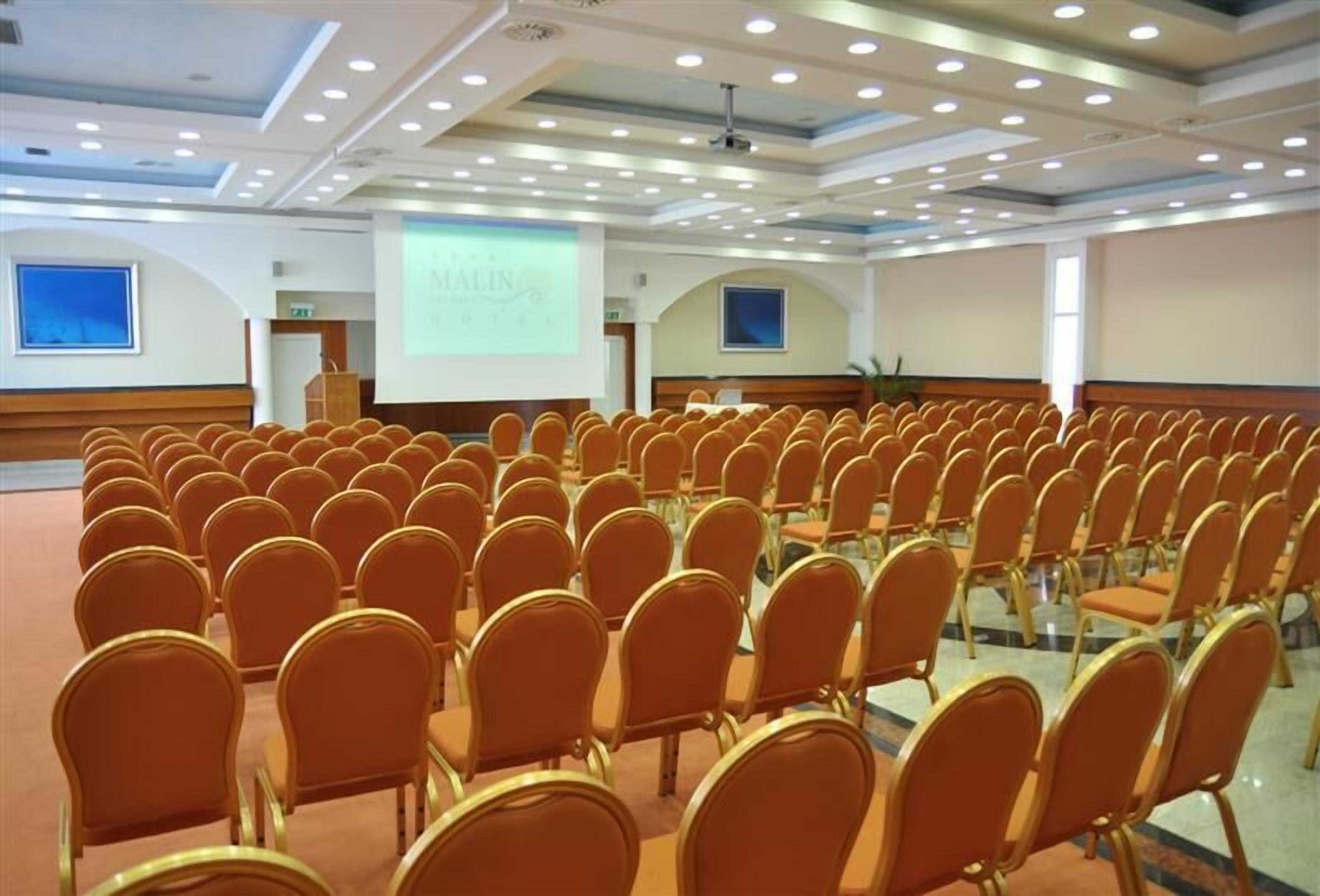Une salle de conférence avec des rangées de chaises orange, un pupitre et un écran de projection affichant le mot « MALIN ».