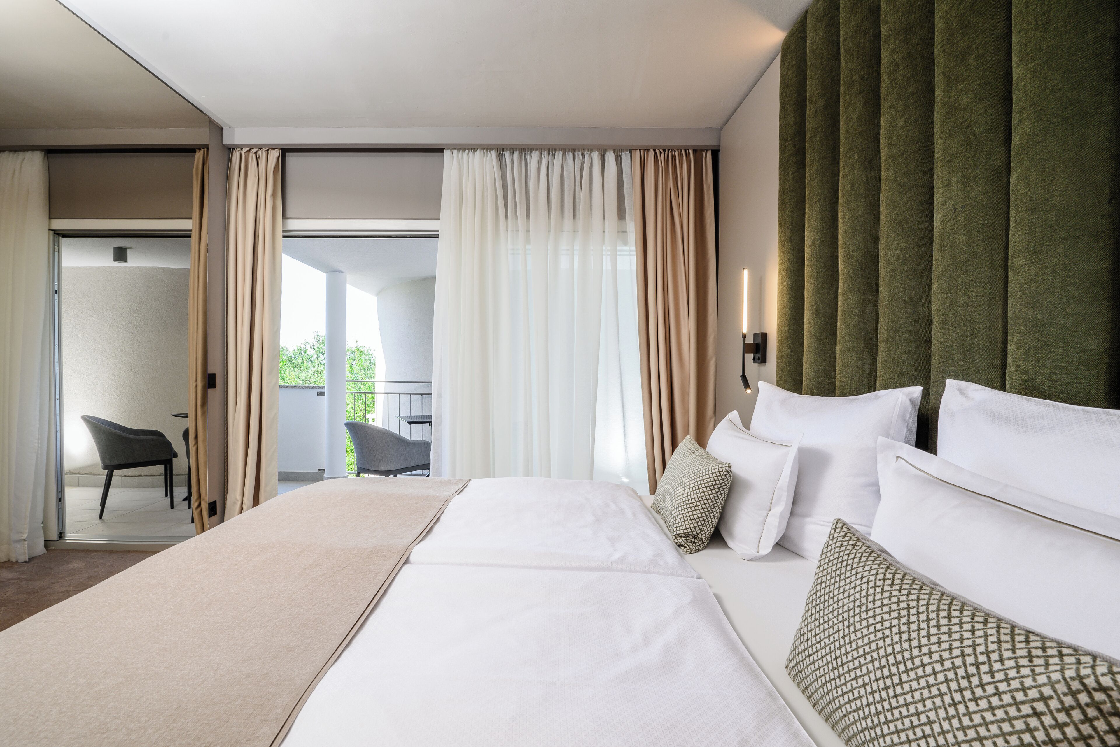 Ein modernes Hotelzimmer mit einem großen Bett, einem grünen gepolsterten Kopfteil und einem Balkon mit einem Sessel.