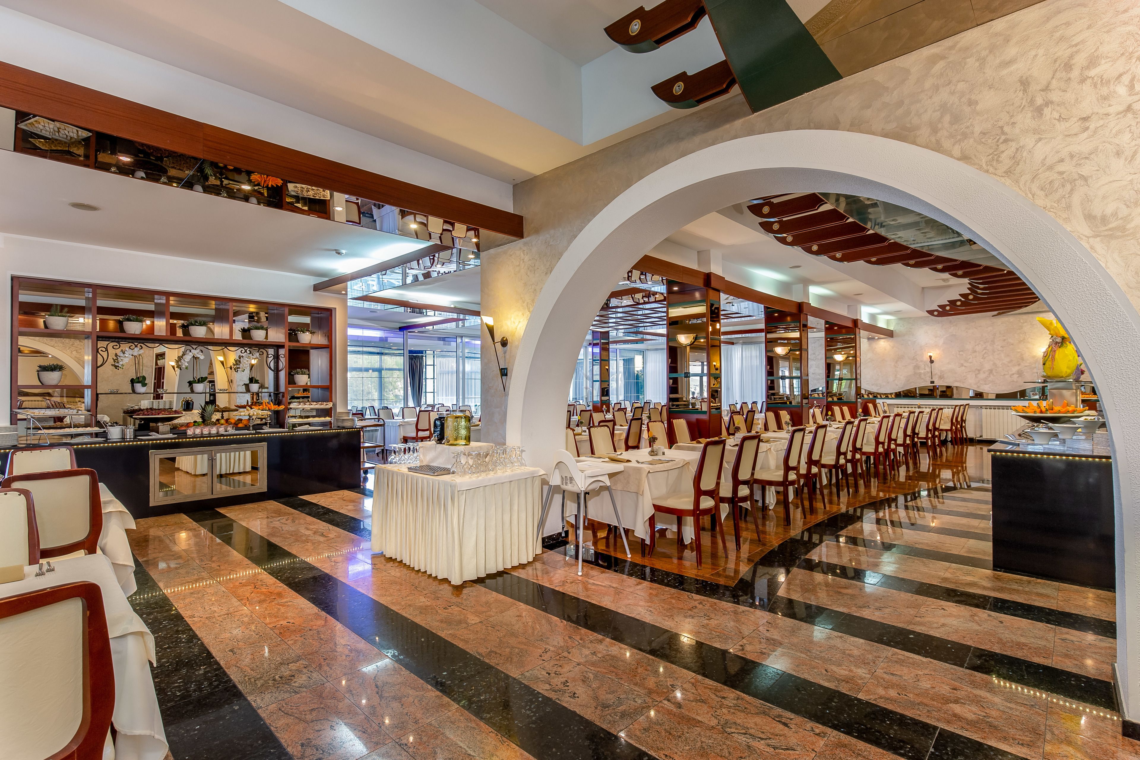 A spacious dining area with a long counter, multiple tables, and chairs, featuring a polished marble floor and a high ceiling with exposed beams.