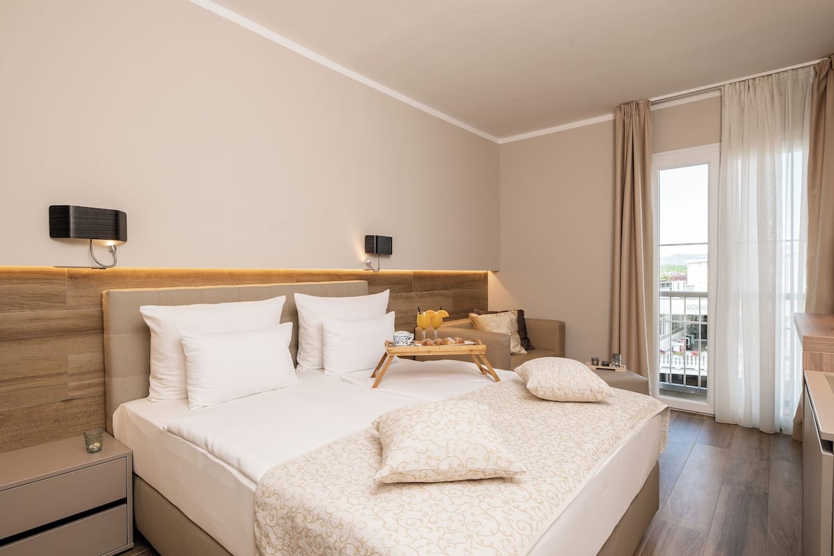 Une chambre d’hôtel moderne dotée d’un grand lit, de tables de chevet, d’une tête de lit en bois, d’un balcon avec des rideaux et offrant une vue sur la ville.