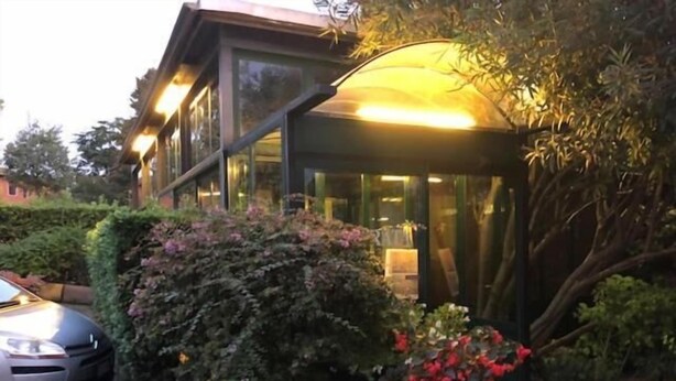 Un edificio moderno con fachada de vidrio, una zona de estar exterior cubierta y un jardín bien cuidado con arbustos y flores.