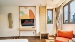 Habitación de hotel moderna con suelo de madera, cama, televisor montado en la pared, una pequeña mesa de madera, una silla naranja y una ventana con cortinas.