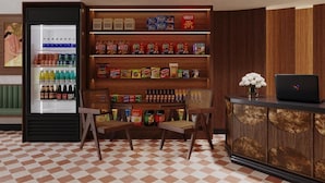 Un hall de hotel con un bar de aperitivos que presenta un refrigerador lleno de bebidas y una estantería con diferentes aperitivos y papas fritas. Hay un ordenador portátil sobre una mesa de madera con un jarrón de flores.
