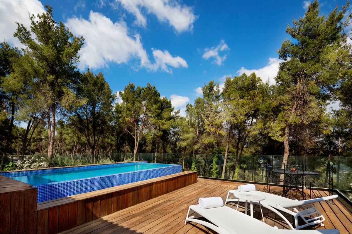 A wooden deck with a swimming pool, lounge chairs, and a glass-enclosed table.