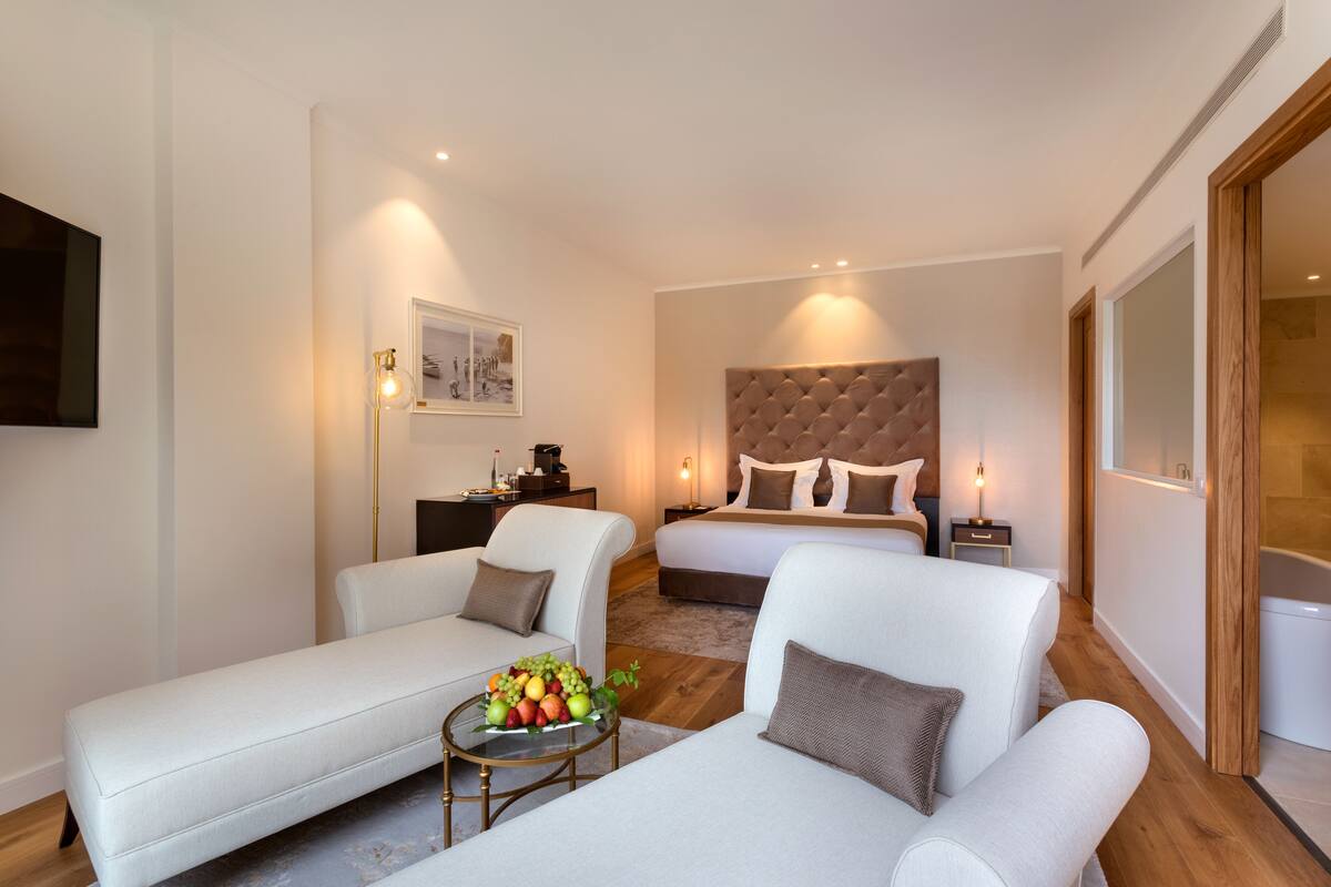 A modern hotel room with a large bed, two white sofas, a small round table with fruit, and a painting on the wall.