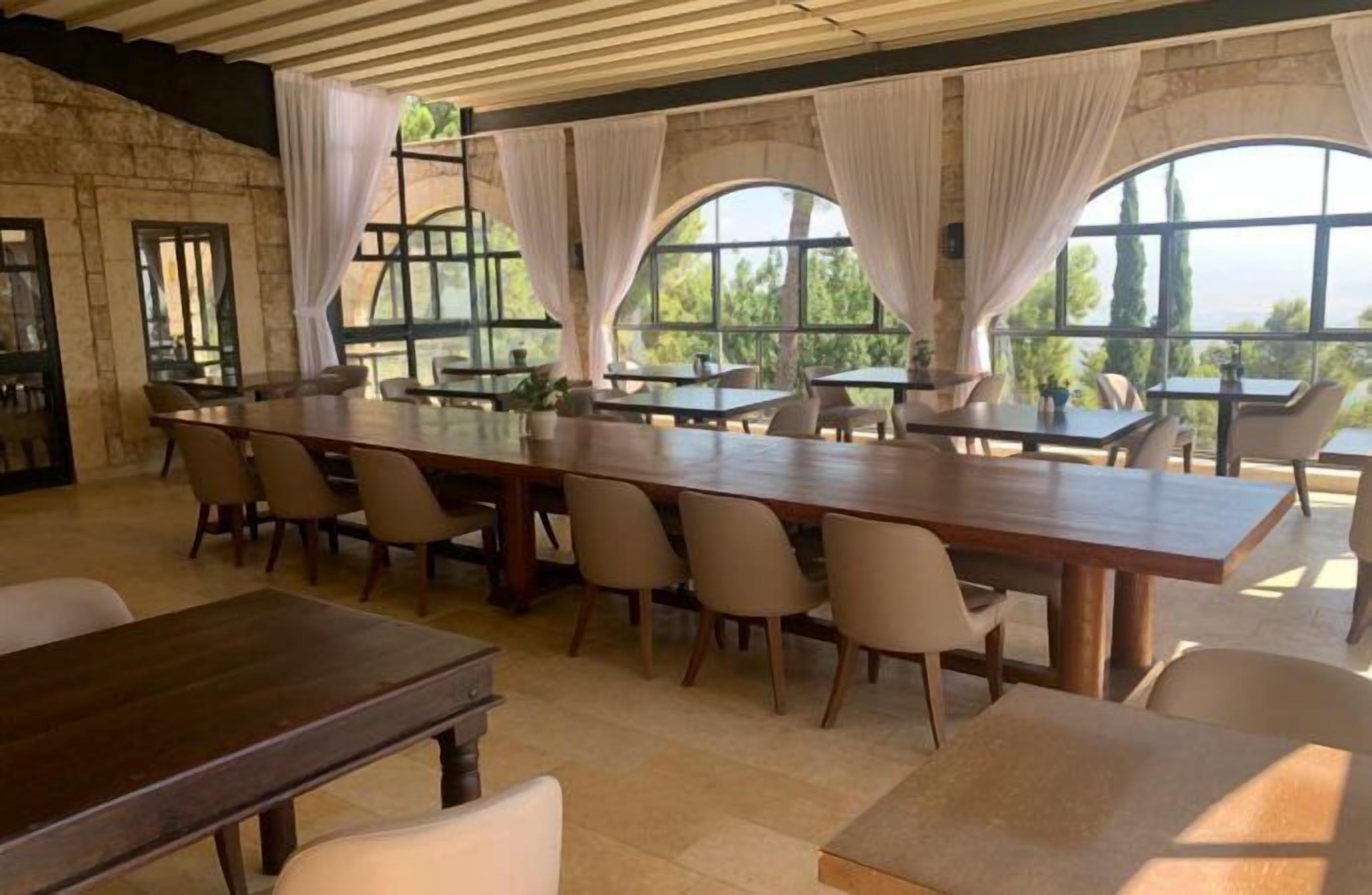 A dining area with a long wooden table and chairs, large windows with curtains, and a stone wall.