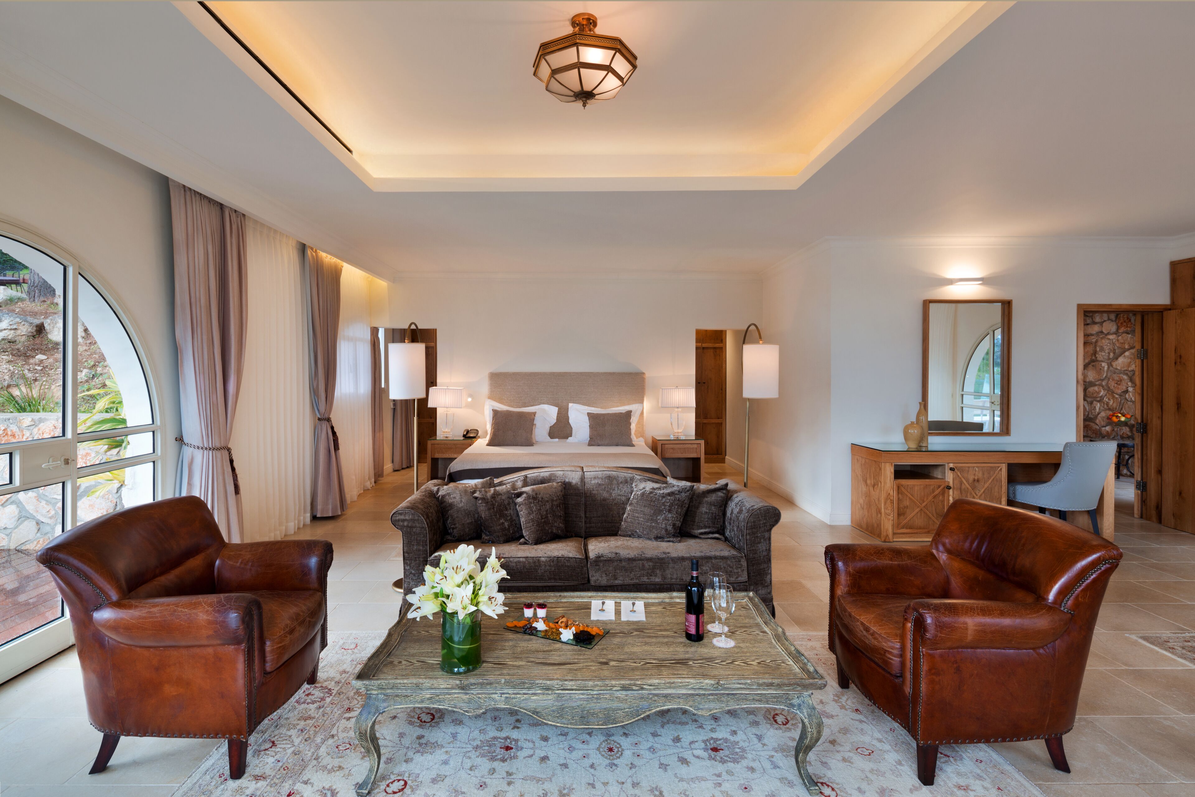A hotel room with a bed, two leather armchairs, a coffee table with a vase of flowers, and a desk with a mirror.