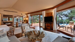 A spacious living room with a glass coffee table, comfortable seating, a flat-screen TV, and a view of a swimming pool and trees.