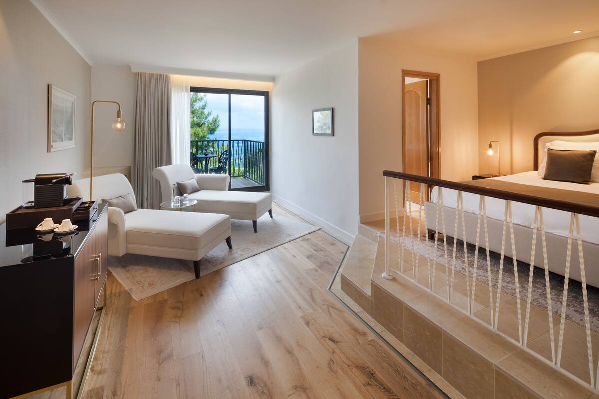 A modern hotel room with a balcony, a sofa, a dining table, and a staircase leading to an upper floor.