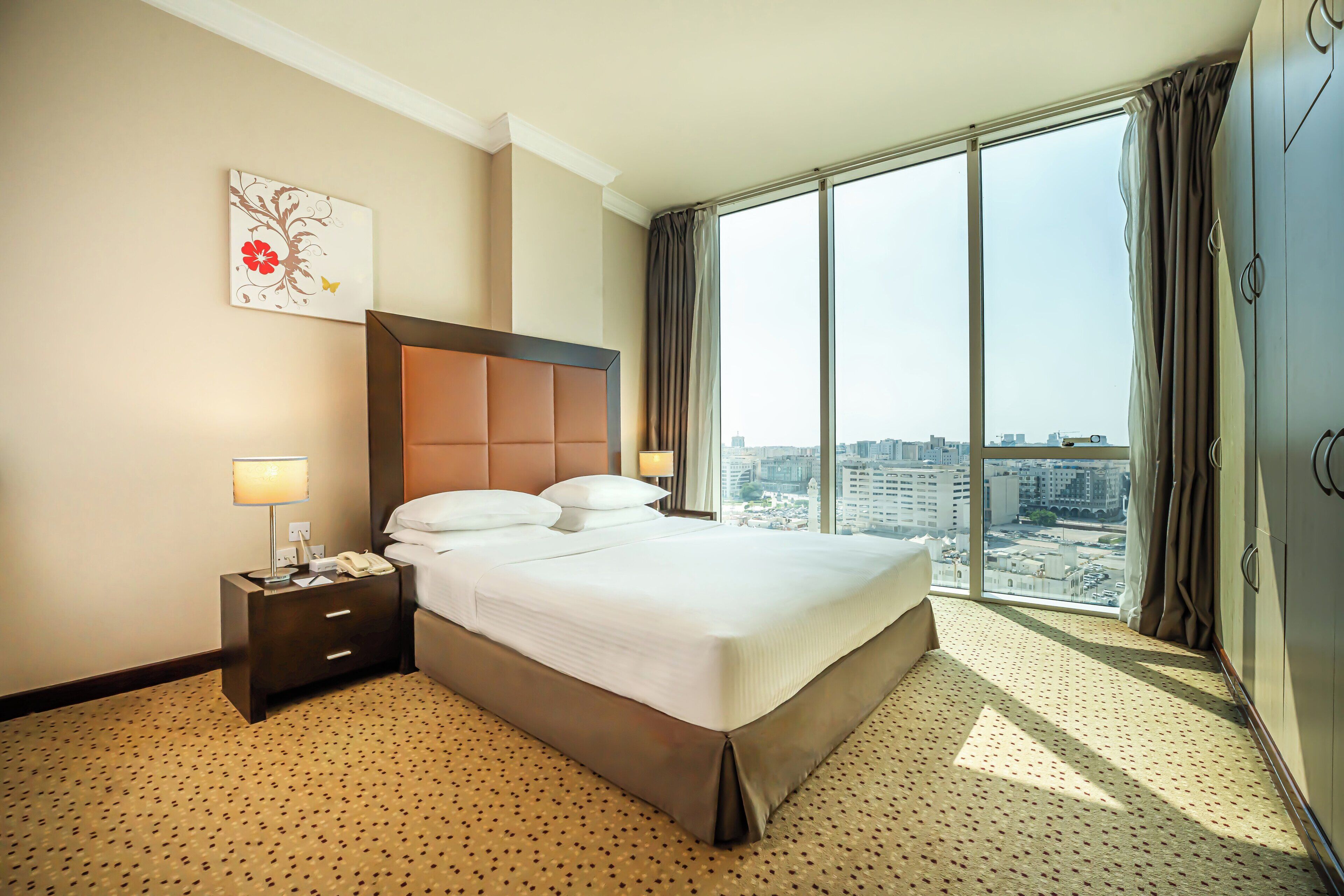 Habitación de hotel con una cama grande, una mesita de noche con lámpara, vista a la ciudad a través de amplias ventanas y un cuadro en la pared.