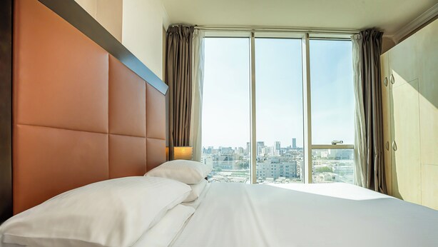 Una habitación de hotel con una cama grande, cabecero, vistas de la ciudad a través de ventanales del suelo al techo y un armario.