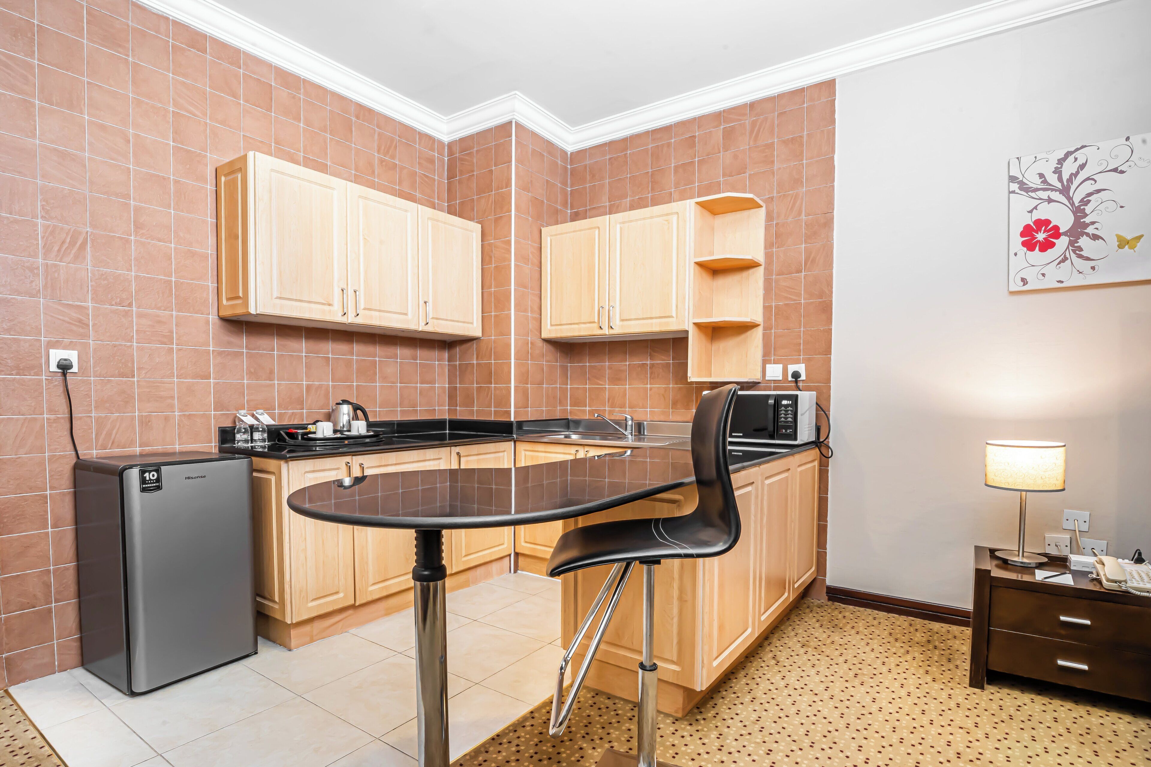 Una cocina compacta con armarios de madera, un refrigerador, un microondas, una mesa de comedor con sillas y un mueble pequeño con una lámpara.