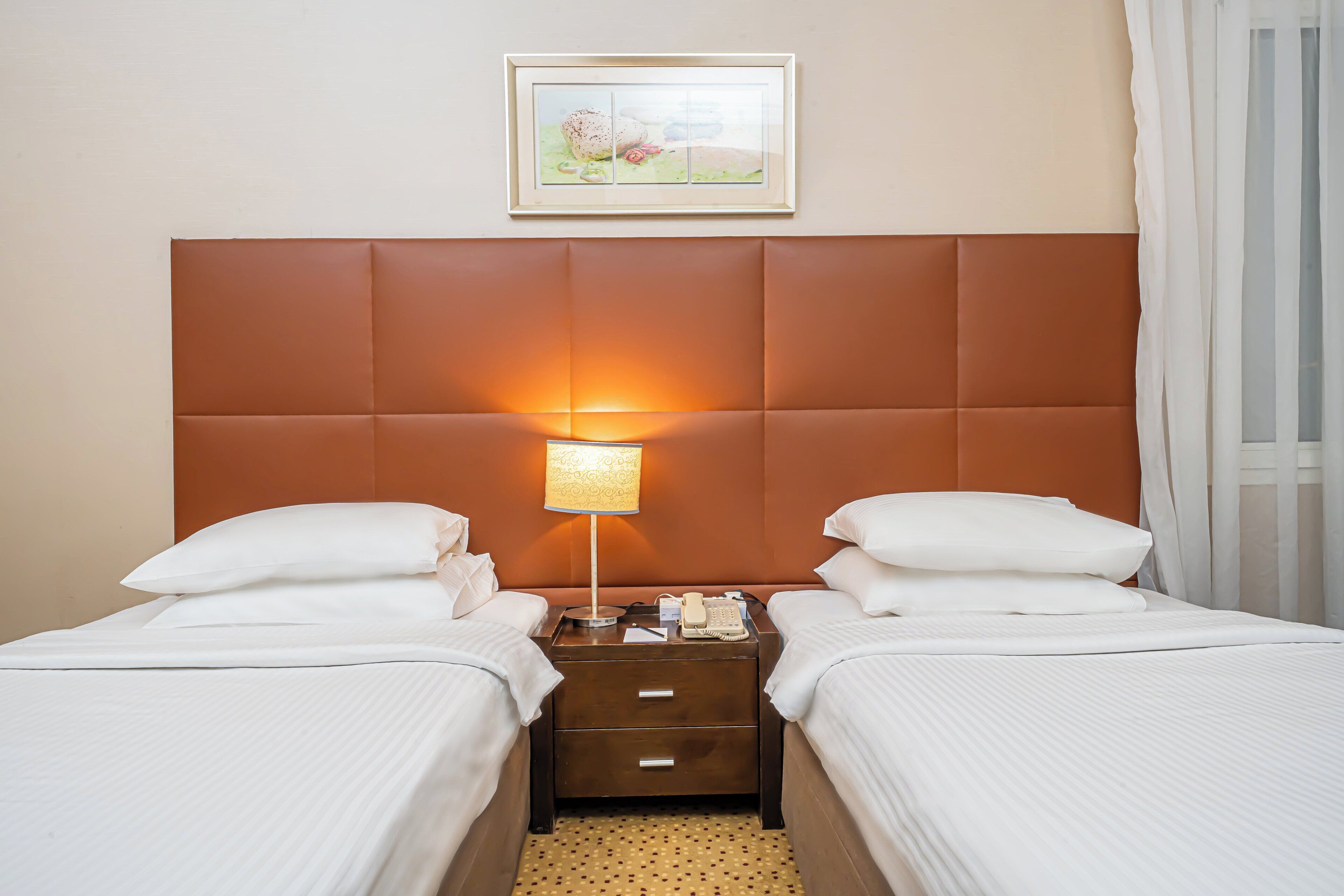 A hotel room with two beds, a nightstand with a lamp, a framed picture on the wall, and a window with curtains.