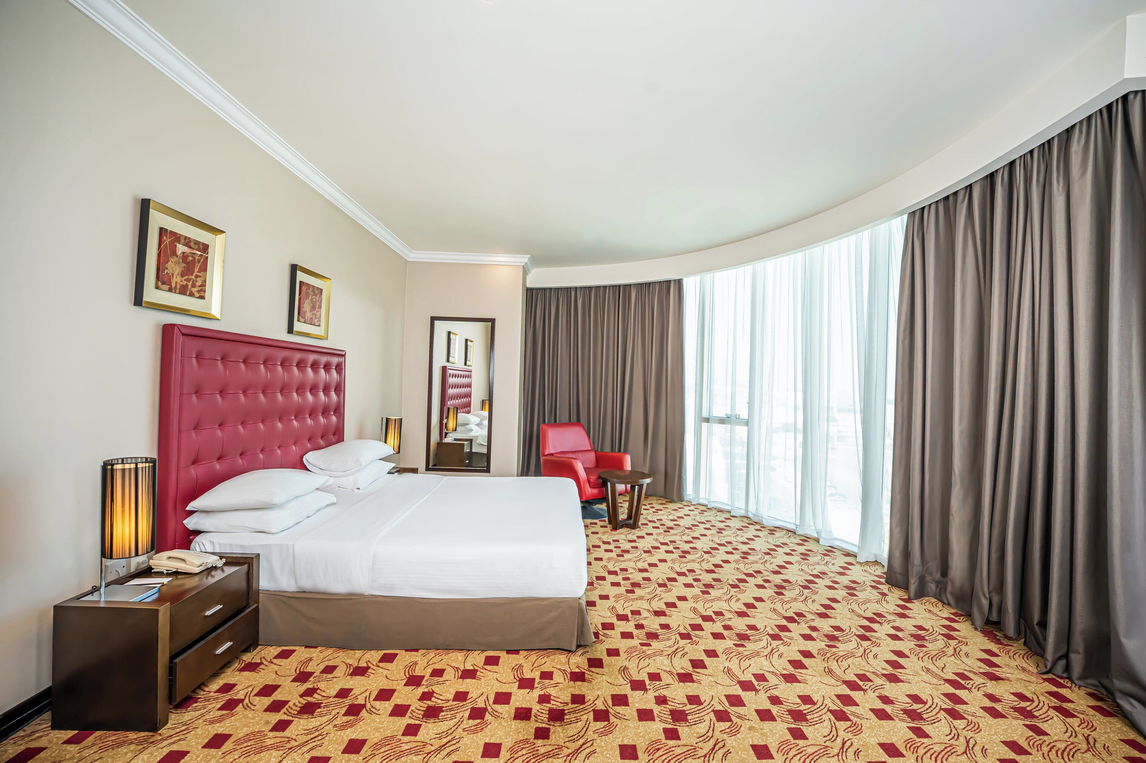 Una camera d'albergo con un letto grande, una testiera rossa, una poltrona rossa, un comodino con una lampada e un'ampia finestra con tende.