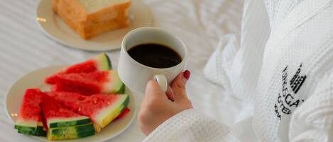 Una persona con bata blanca sosteniendo una taza de café, con un plato de sandía en rodajas y una rebanada de tostada sobre una cama.