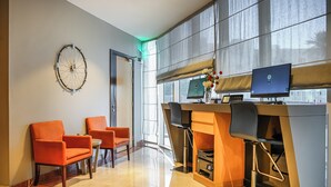 Un vestíbulo moderno de hotel con mostrador de recepción, dos sillas naranjas, un reloj de pared grande y un ventanal con vistas.