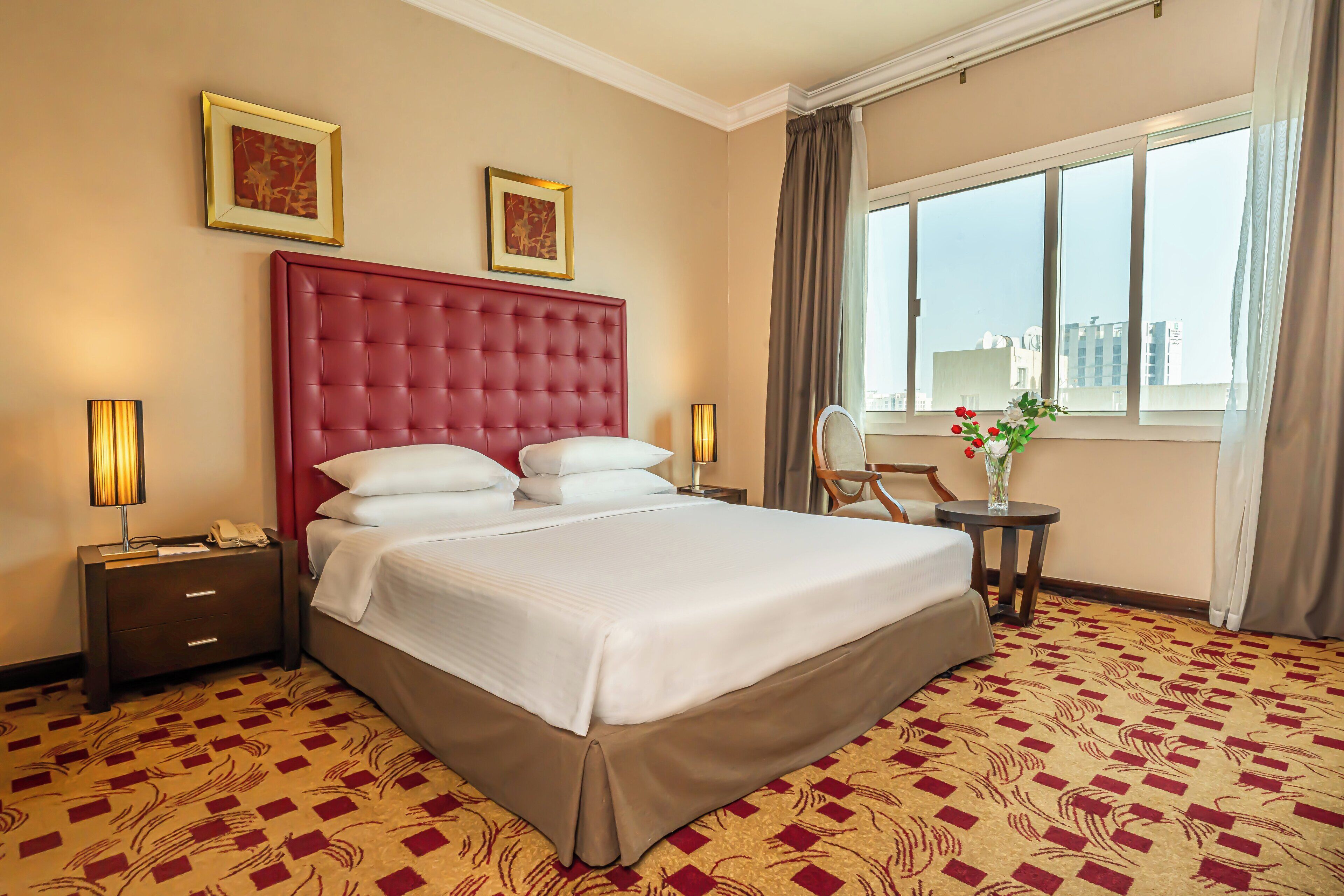Habitación de hotel con una cama grande, dos mesitas de noche, una silla, una mesita con un florero y una ventana con cortinas.