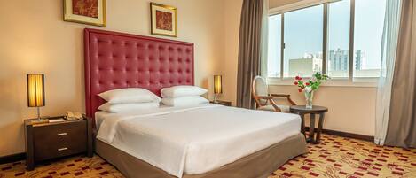 Habitación de hotel con una cama grande, dos mesitas de noche, una silla, una mesita con un florero y una ventana con cortinas.