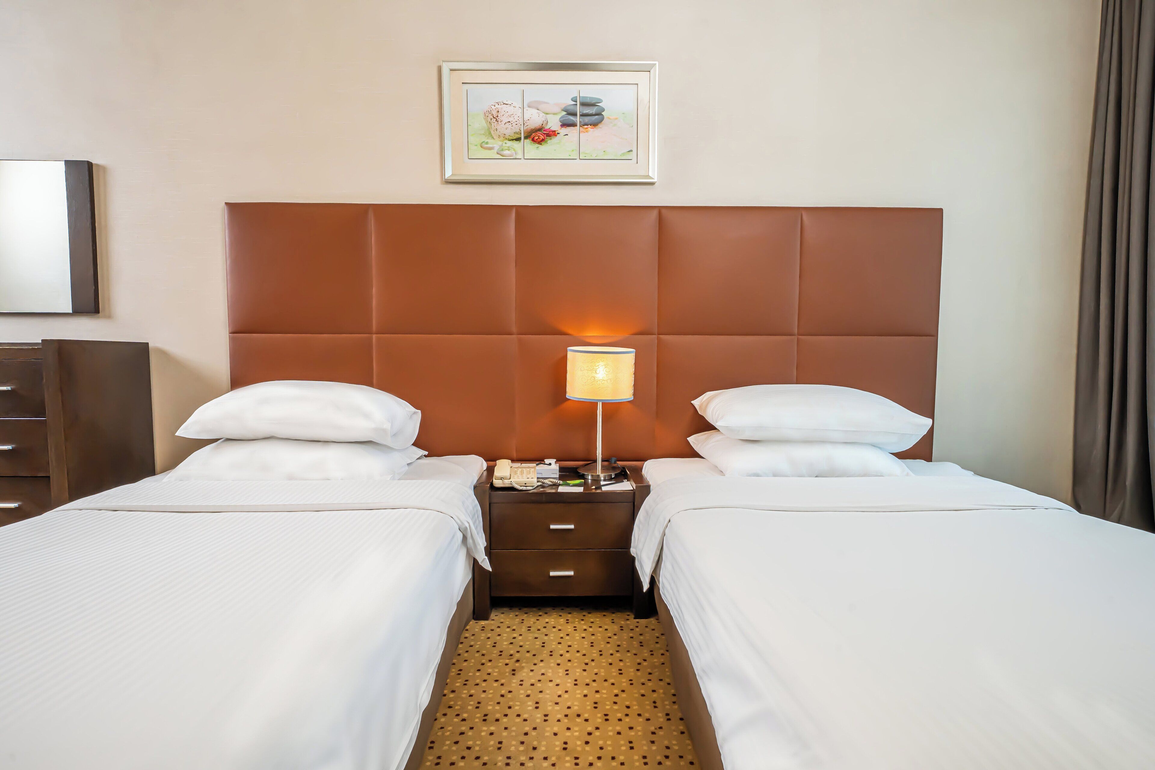 Habitación de hotel con dos camas, una mesita de noche con lámpara, un cuadro enmarcado en la pared y alfombra marrón.