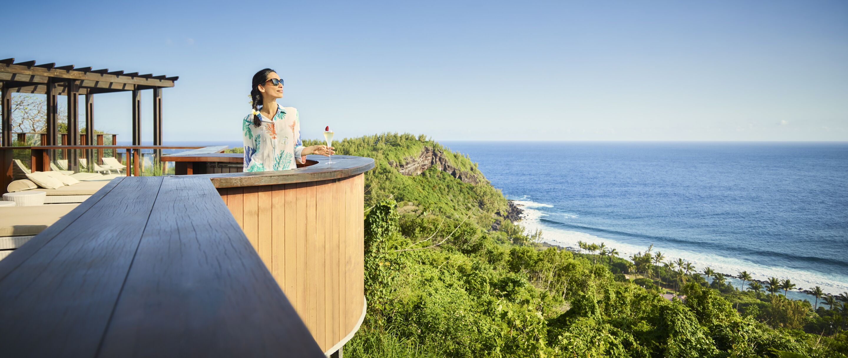 A person standing on a wooden deck overlooking a coastal landscape with a clear view of the ocean and a lush green hillside.