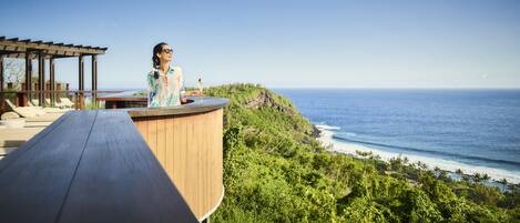 A person standing on a wooden deck overlooking a coastal landscape with a clear view of the ocean and a lush green hillside.