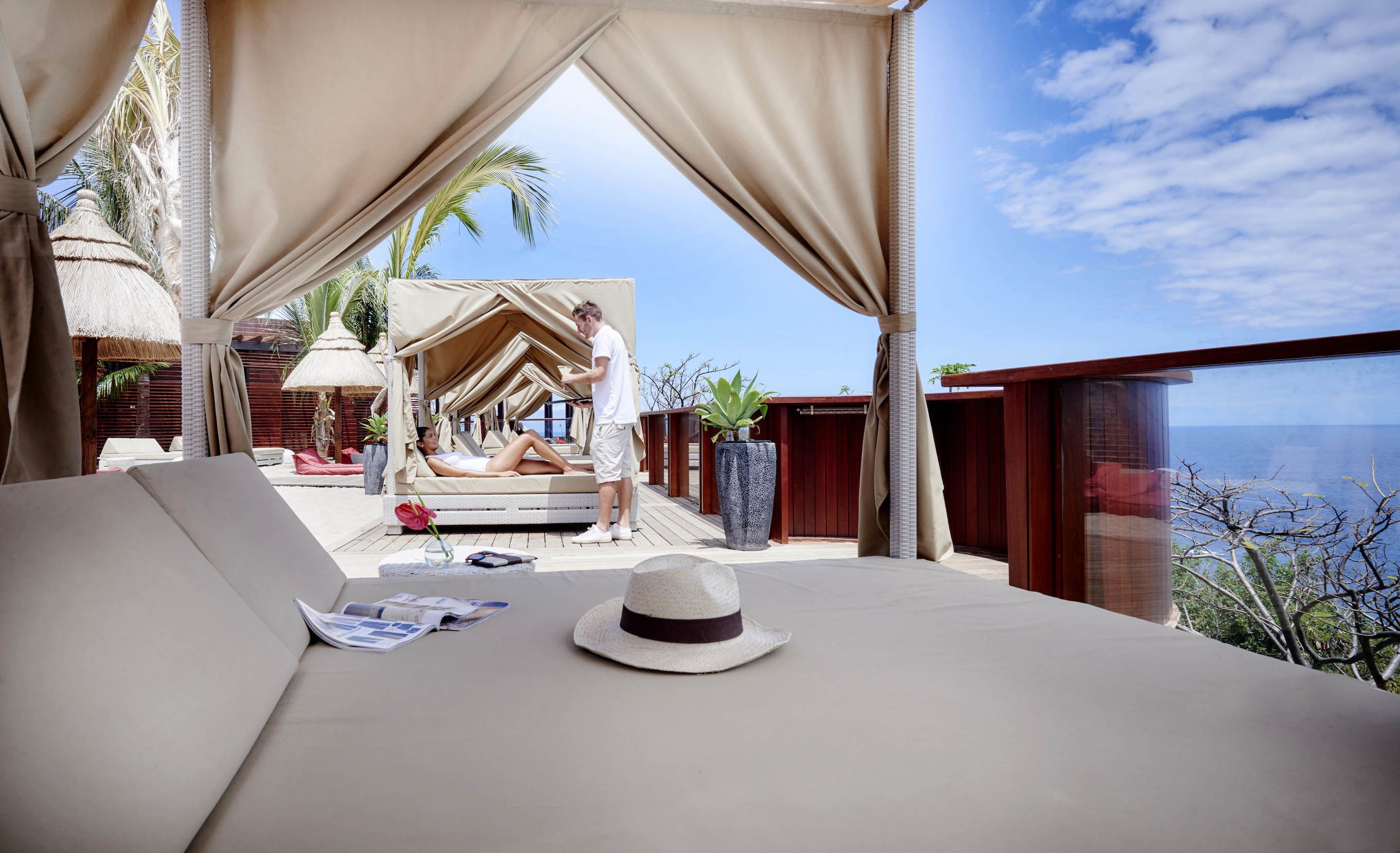 A beachside lounge with a view of the sea, a thatched-roof structure, and a large potted plant.