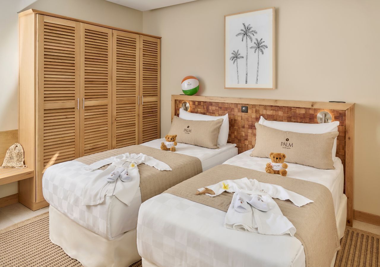 A hotel room with two beds, a wooden wardrobe, a framed picture of palm trees, and a small ball on the wall.