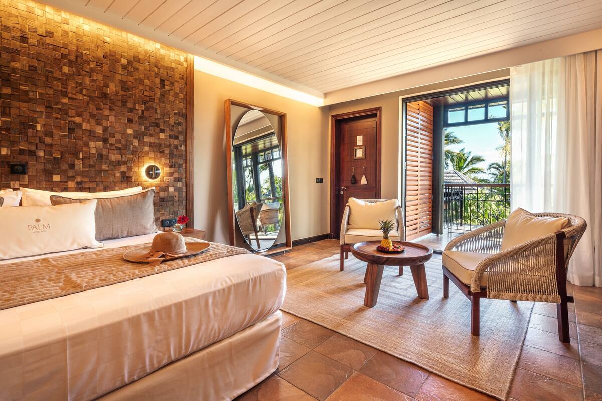 A hotel room with a large bed, a sitting area with a wicker chair, a small table, and a balcony with a view of palm trees.