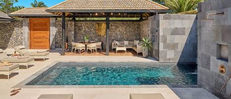 Un área de piscina con sillones de descanso, una zona de estar cubierta y un muro de piedra.