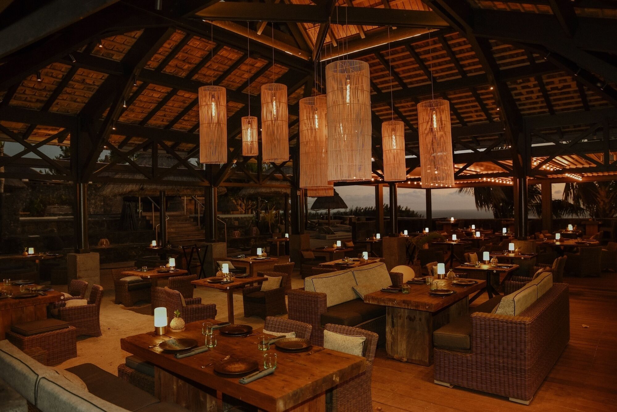 Restaurante com altos tetos de madeira, lustres de vime suspensos e mobiliário de vime. Mesas montadas com pratos, copos e velas.