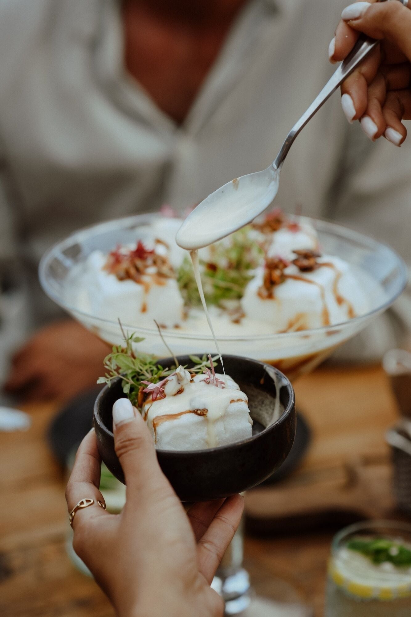 A person serving a dessert with a spoon, garnished with herbs and sauce, in a clear bowl.