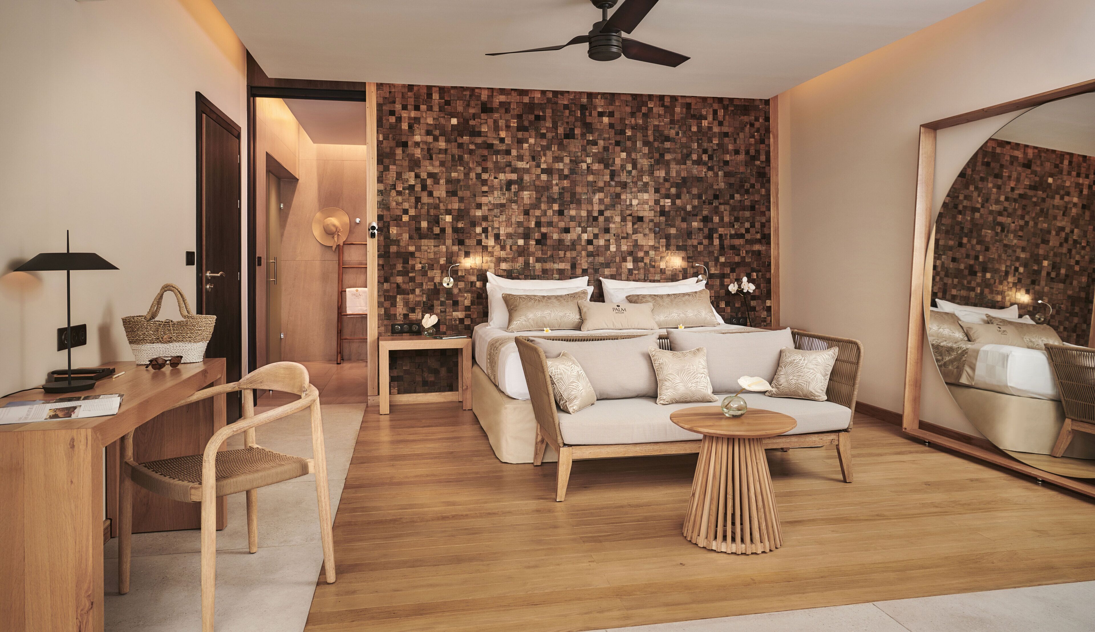 Quarto moderno com piso de madeira, cama com várias almofadas, sofá, mesa redonda pequena, mesa de cabeceira, abajur, cadeira, espelho grande e ventilador de teto.