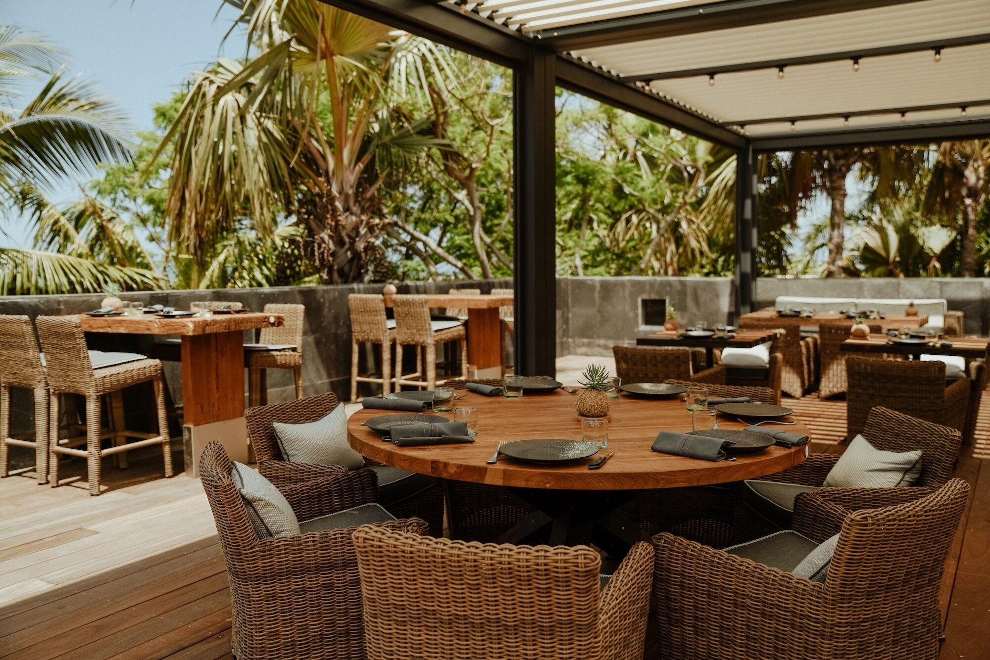 An outdoor dining area with wicker furniture and a wooden table, surrounded by lush greenery and palm trees.