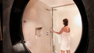 A person in a white bathrobe standing in a glass-enclosed shower area with a built-in shelf holding toiletries.