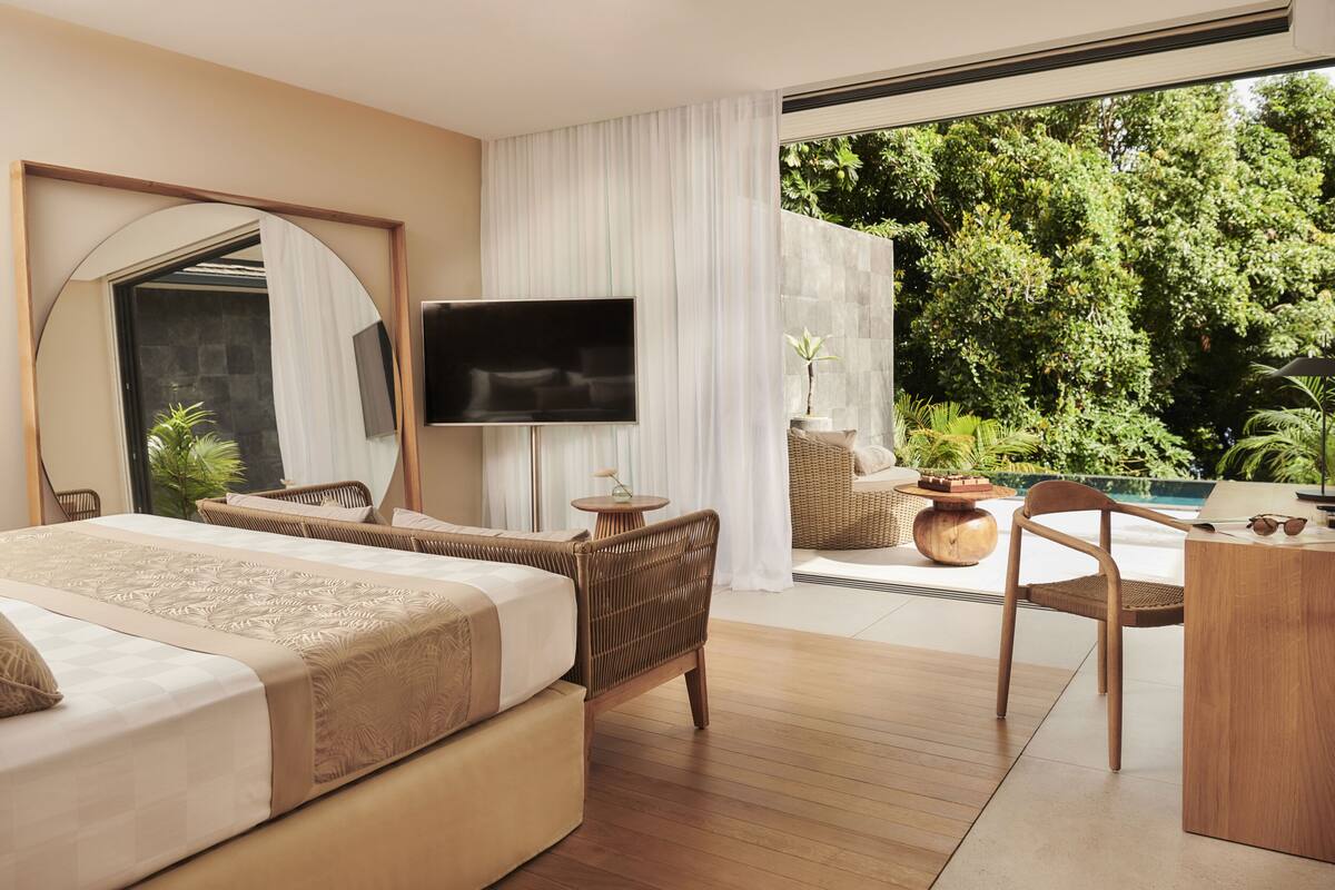 A modern hotel room with a large bed, a television, a dining area, and a view of lush greenery.