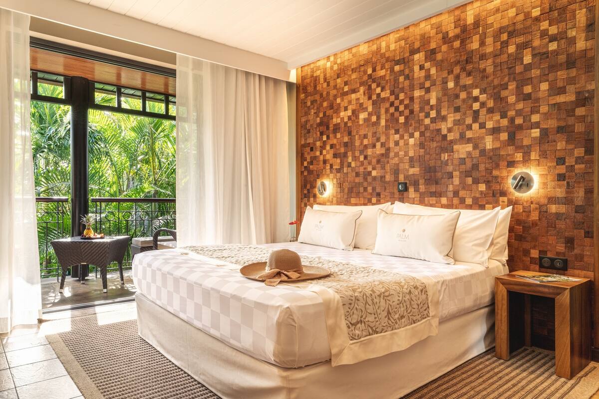 A spacious bedroom with a large bed, a wooden accent wall, a small table, and a balcony with a view of lush greenery.