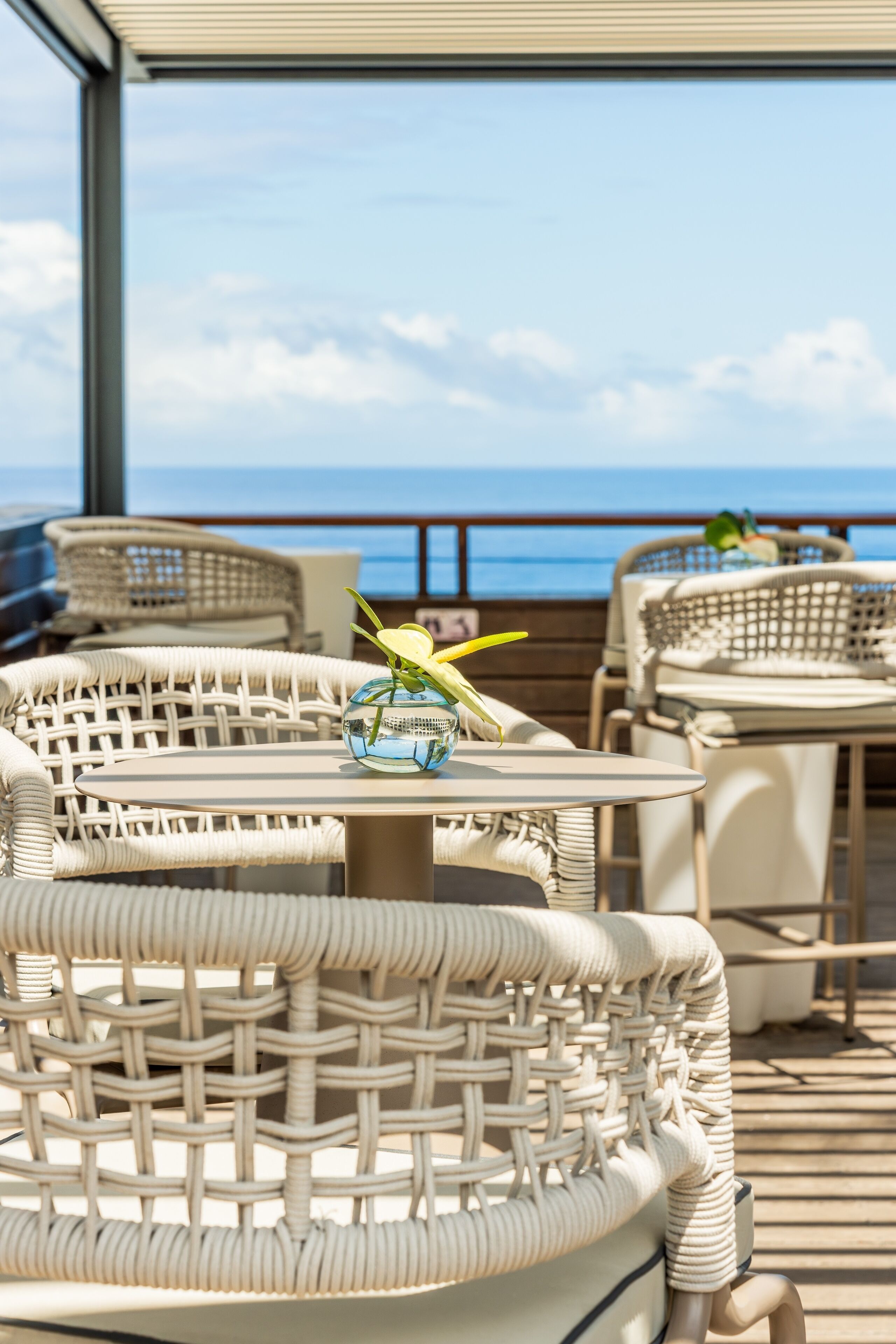 Ein Balkon mit Korbmöbeln, einem Glastisch und einem Dekorationsgegenstand, mit Blick auf das Meer.
