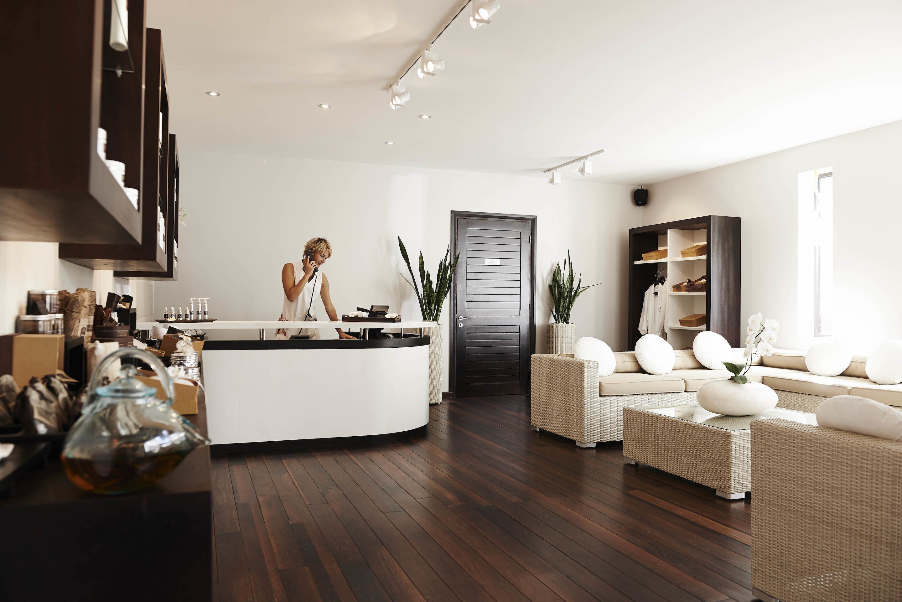 A modern living room with a white reception desk, dark wooden flooring, wicker furniture, and a dark door.