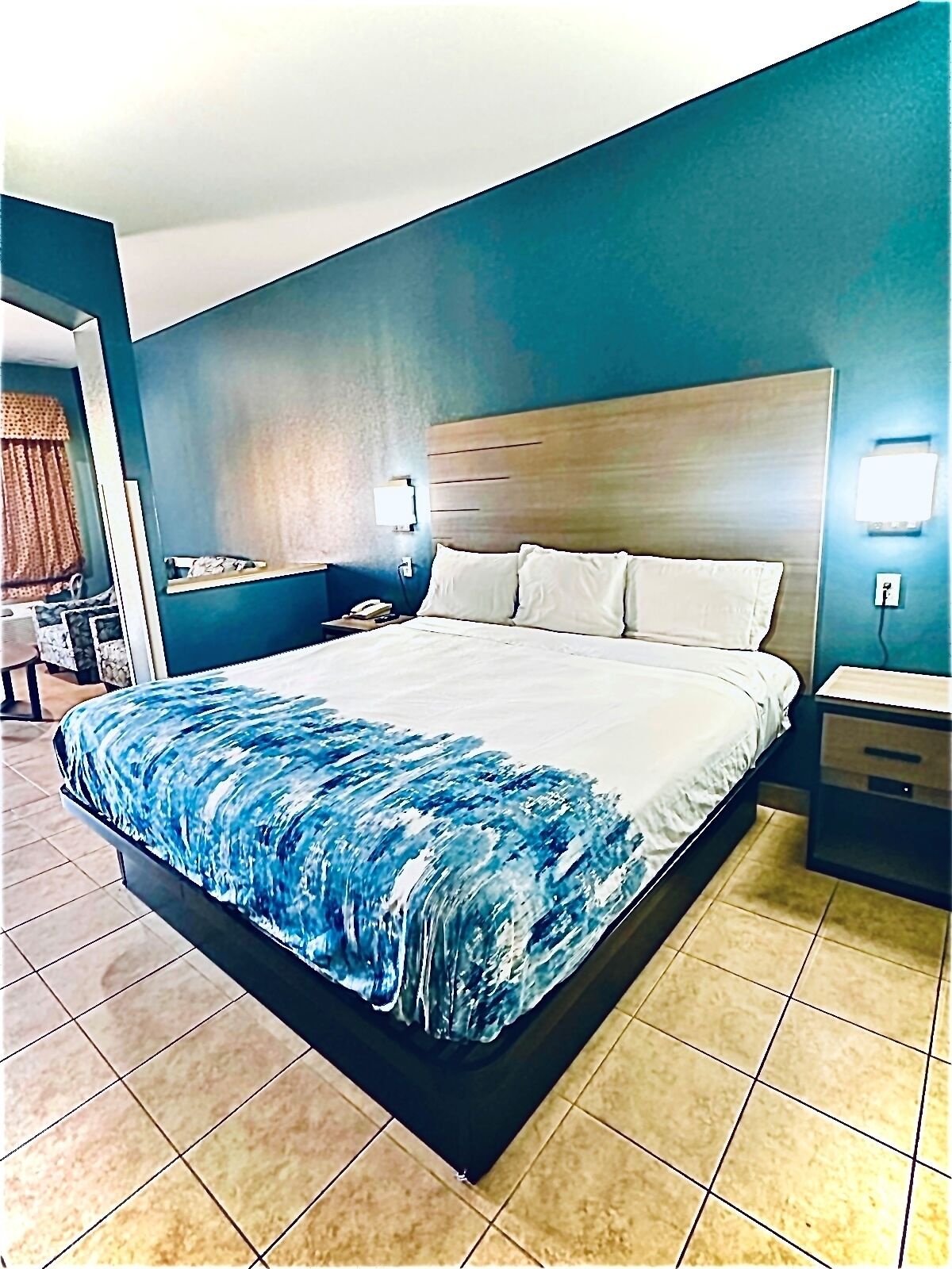 A hotel room with a large bed, a wooden headboard, a nightstand, a bedside table, and a wall-mounted lamp.