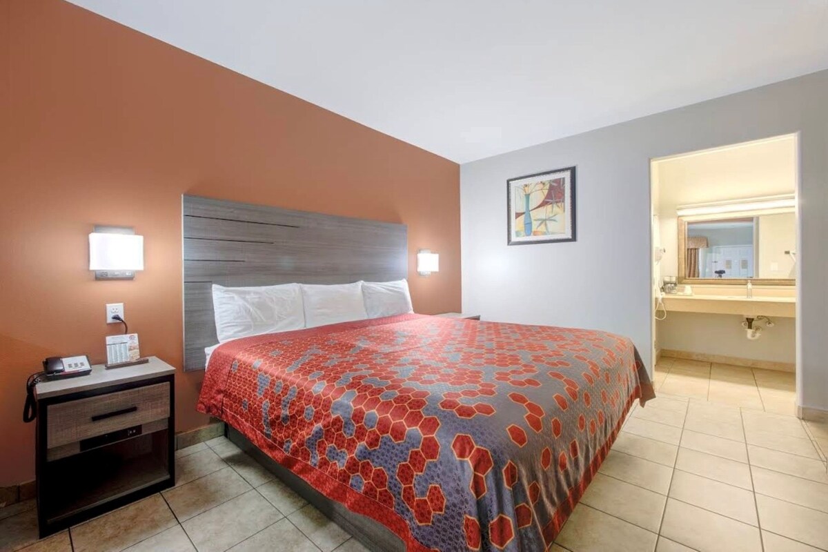A hotel room with a bed, bedside table, wall-mounted lights, a painting, and a bathroom with a sink and mirror.